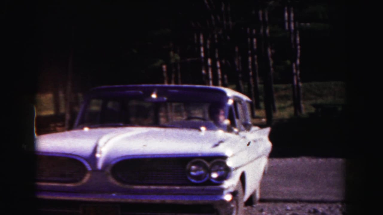 White 1960s sedan parked on a gravel area near trees.