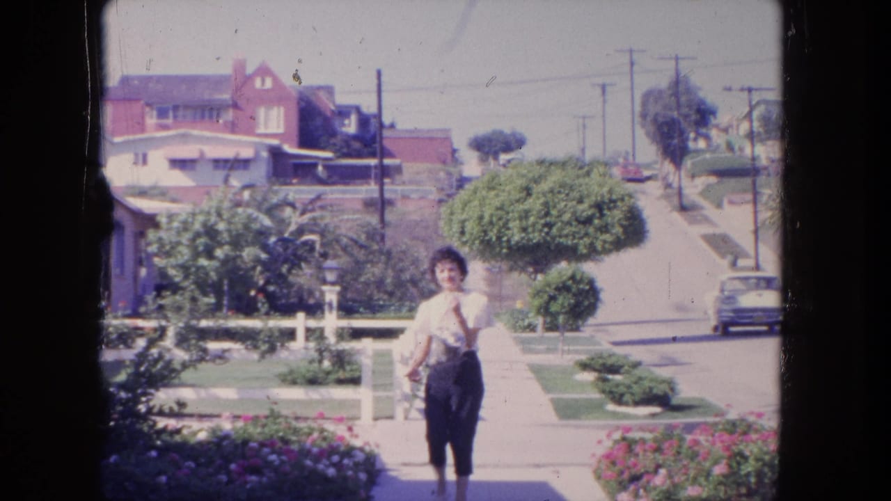 In 1962 Tijuana, a woman walks toward the camera along a suburban sidewalk with lawns, houses, and utility poles behind her.