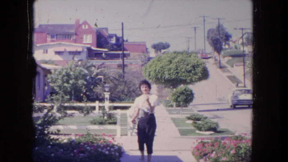 In 1962 Tijuana, a woman walks toward the camera along a suburban sidewalk with lawns, houses, and utility poles behind her.