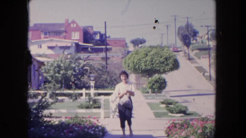 A woman in a light blouse and dark pants faces the camera on a Tijuana residential sidewalk, with houses and planted yards in view.