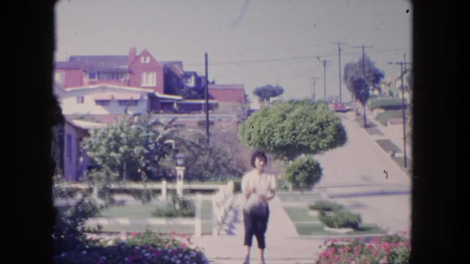 The woman continues along the sidewalk in a 1962 Tijuana residential setting, seen against lawns, shrubs, houses, and overhead utility lines.