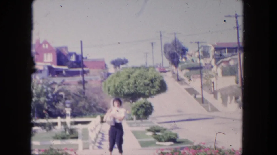 A woman stands or walks near the lower center of the frame on a Tijuana sidewalk, with mid-century houses and a sloped street behind her.