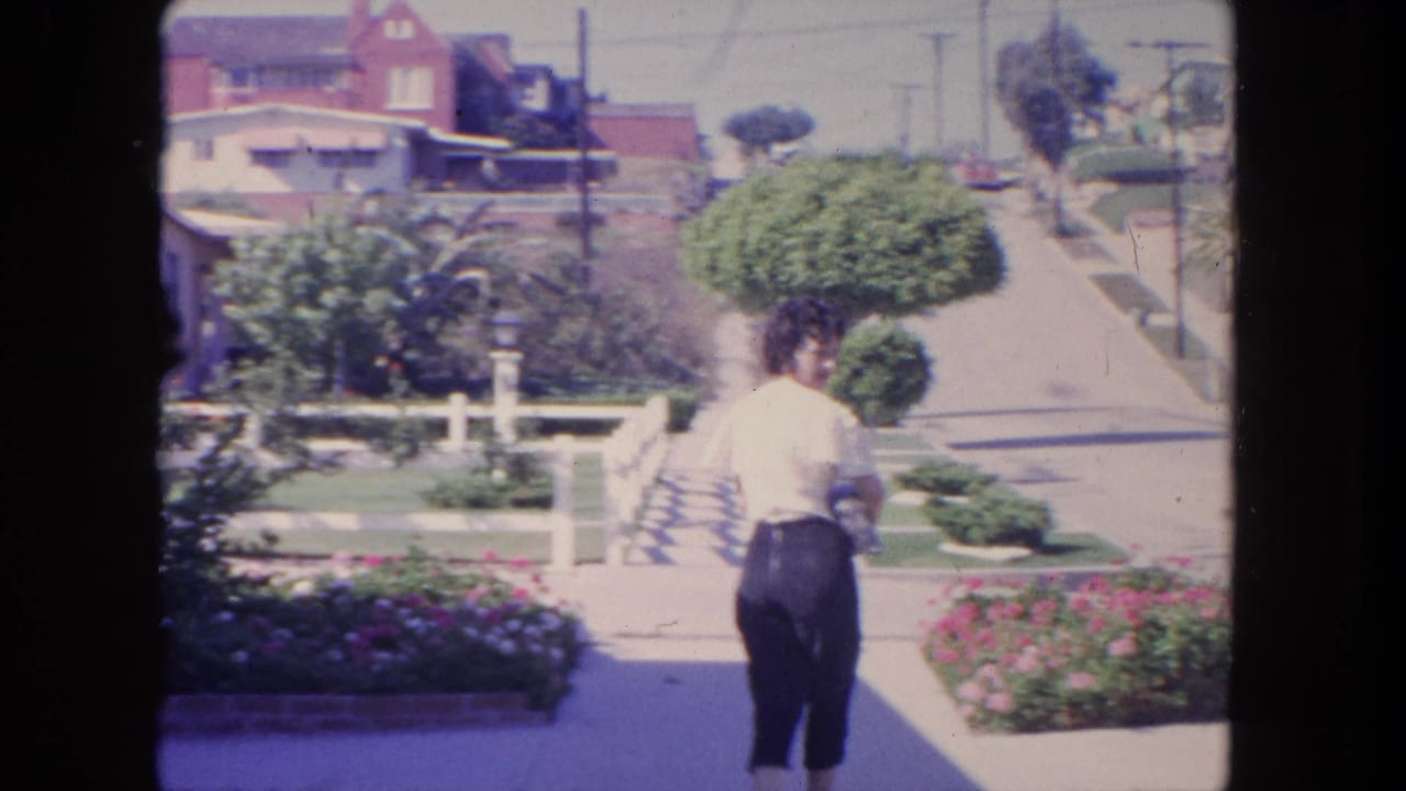 In 1962 Tijuana, the woman turns partway toward the camera beside a planted sidewalk and residential street.