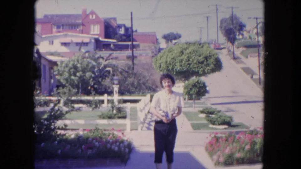 In 1962 Tijuana, the woman faces the home-movie camera on a sunny sidewalk in a residential neighborhood.
