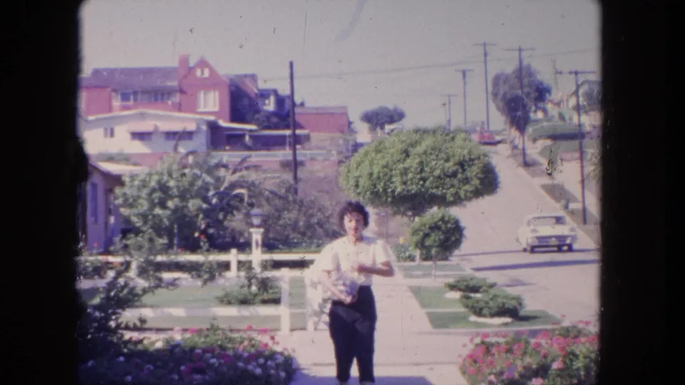 In 1962 Tijuana, the woman remains near the camera while a period car moves along the street in the background.