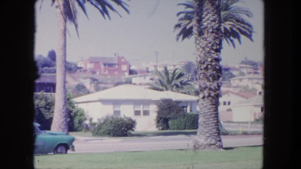 1962 Tijuana home-movie frame showing palm trees, low houses, a street, lawn, and a parked turquoise car.