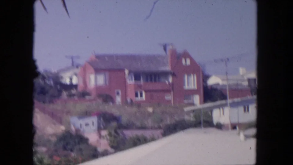 1962 Tijuana frame with a red brick hillside house, nearby residences, utility poles, pale roofline, and palm foliage.