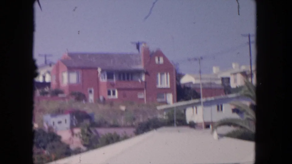 1962 Tijuana home-movie frame centered on a red brick hillside residence, with palms, shrubs, rooftops, and utility poles around it.