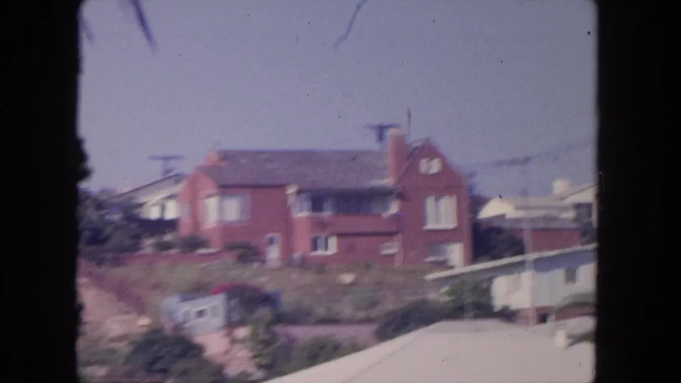 1962 Tijuana frame showing the red brick hillside house beside a pale foreground roof, utility lines, palms, and neighboring homes.