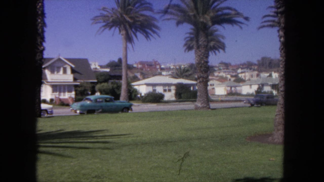 1962 8mm home-movie frame from Tijuana showing a blue car by grass, palm trees, road, and low residential buildings.