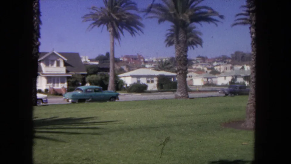 1962 8mm home-movie frame from Tijuana showing a blue car by grass, palm trees, road, and low residential buildings.