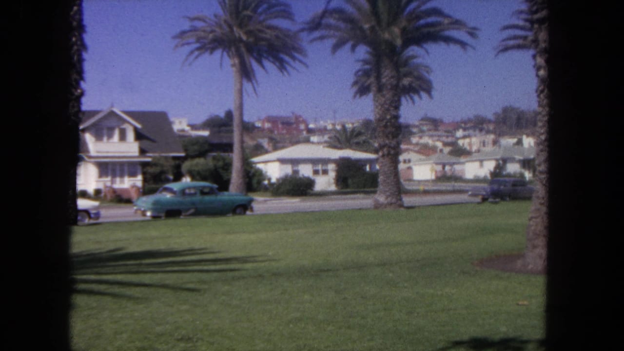 1962 Tijuana street view with a blue car, palm trunks, grass, and residential buildings seen through 8mm film borders.