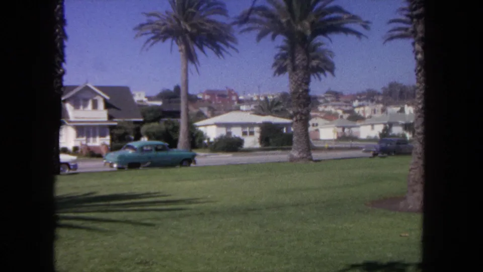 1962 Tijuana street view with a blue car, palm trunks, grass, and residential buildings seen through 8mm film borders.
