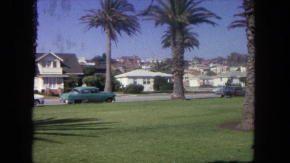 1962 Tijuana home-movie frame showing palm trees, mid-century houses, a lawn, and a turquoise car on a residential street.