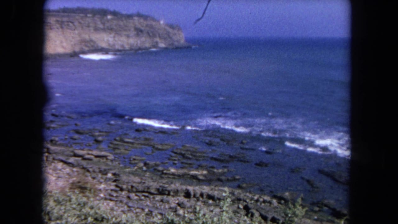 Blue ocean waves breaking over a rocky shoreline with a distant cliff and dark frame edges.