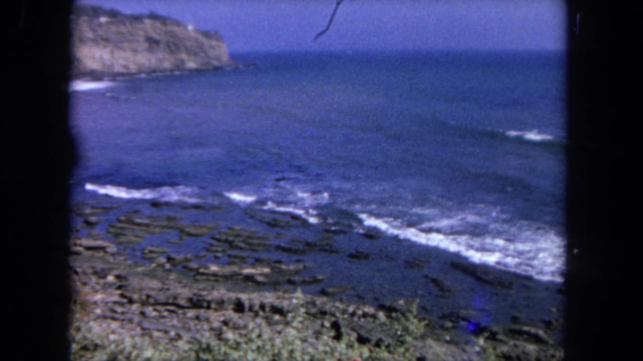 Rocky coast and blue water with white surf lines, framed by dark vertical borders.