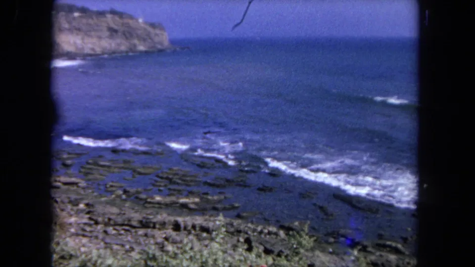 1962 Tijuana-area coastal view with blue water, white wave foam, rocky shelves, and a cliff at the left edge of the frame.