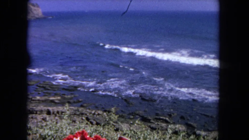 1962 Tijuana-area home-movie frame of white waves spreading across rocky shore, with blue ocean beyond and vegetation along the bottom edge.