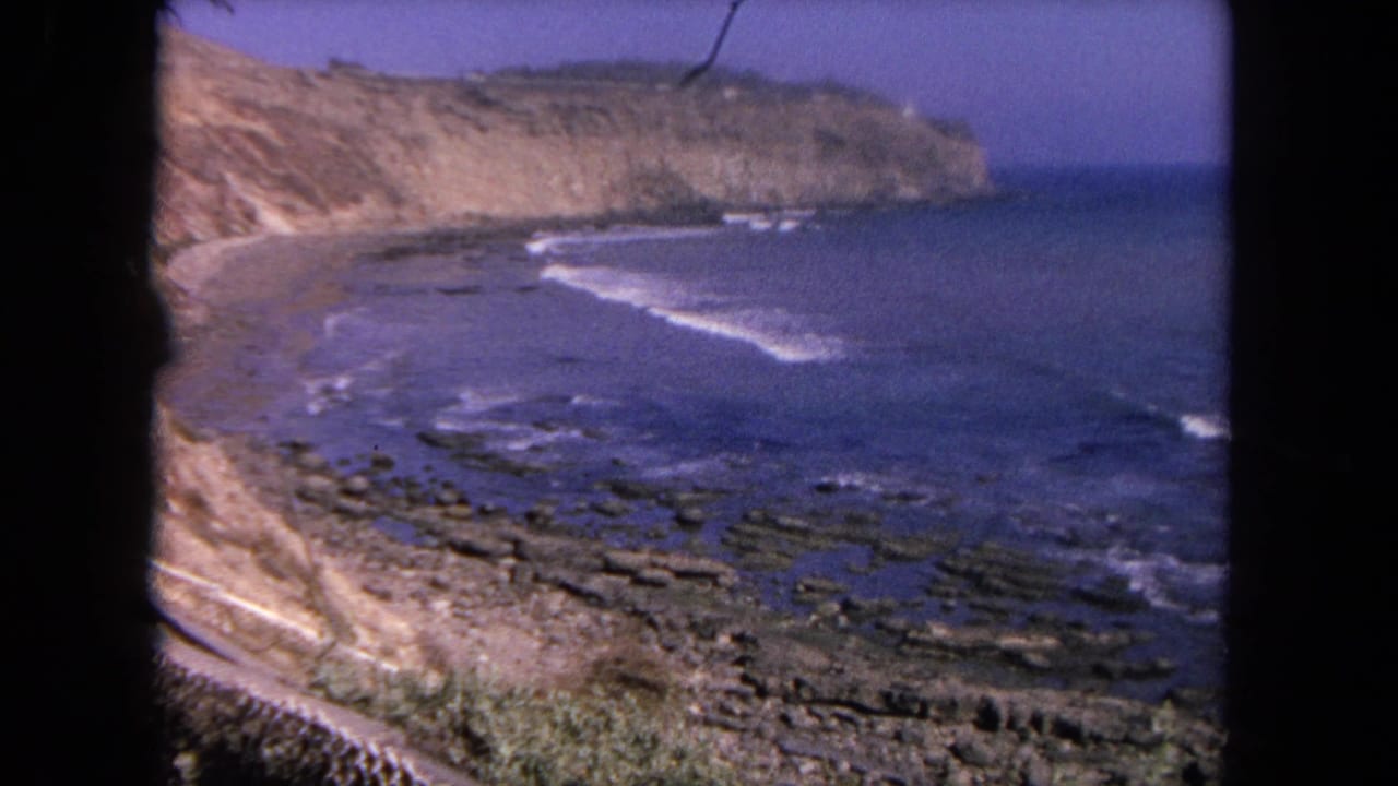 1962 Tijuana coastal view on 8mm film showing waves breaking along a rocky shore beneath tan cliffs and blue sky.