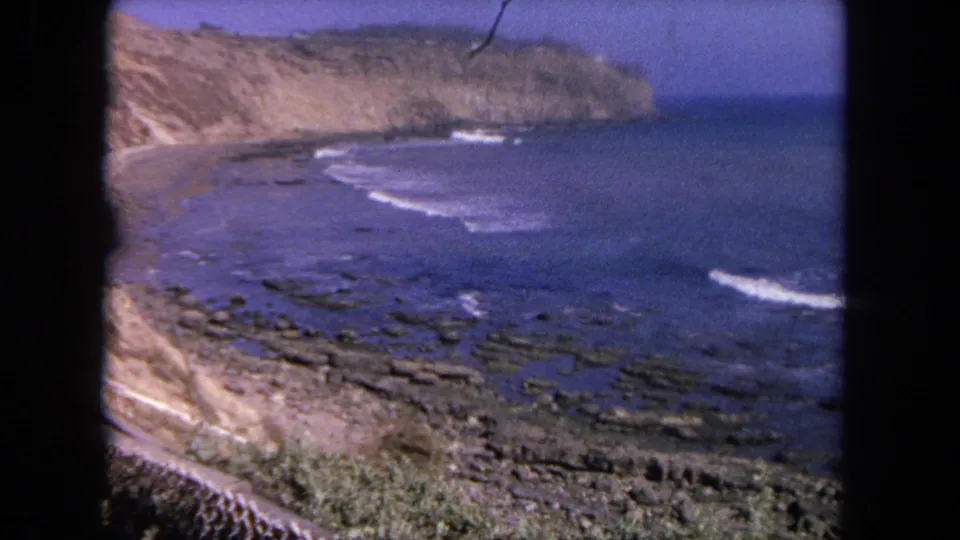 1962 Tijuana shoreline footage showing a cliff face, rocky beach, and white surf lines across saturated blue water.