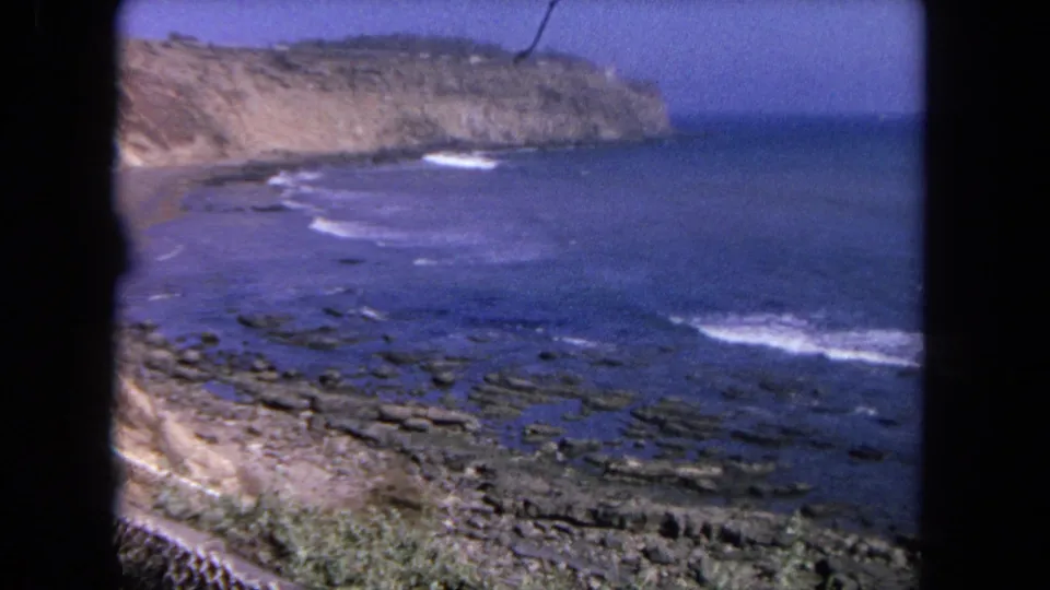 8mm home-movie frame from Tijuana in 1962, looking over blue ocean, rocky shallows, and a tan coastal bluff.