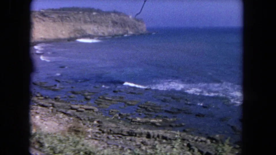 1962 Tijuana coastal scene on 8mm film, with low surf over rocks, open blue water, and a distant tan cliff.