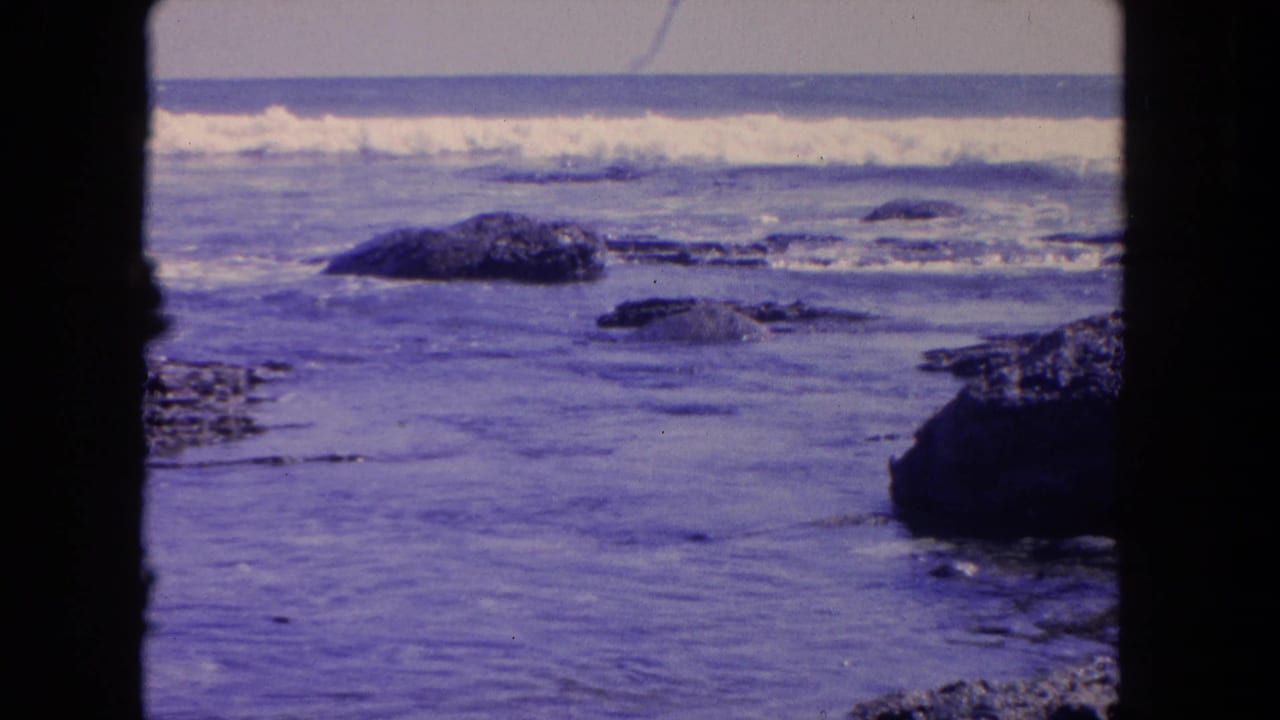Waves roll over dark rocks along a purple-blue shoreline, framed by black vertical film borders.
