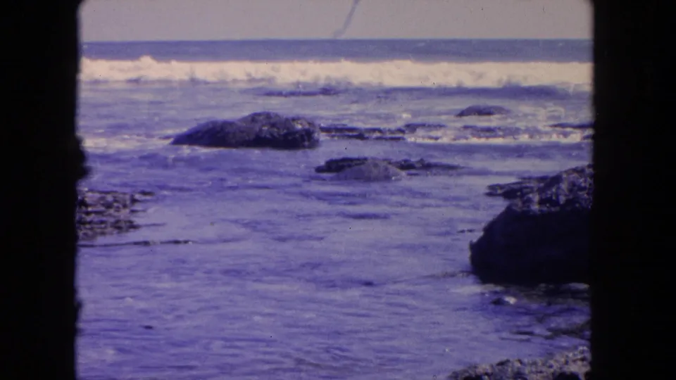 1962 Tijuana coastal footage showing white surf moving over dark boulders in shallow water, with black film-frame edges visible.