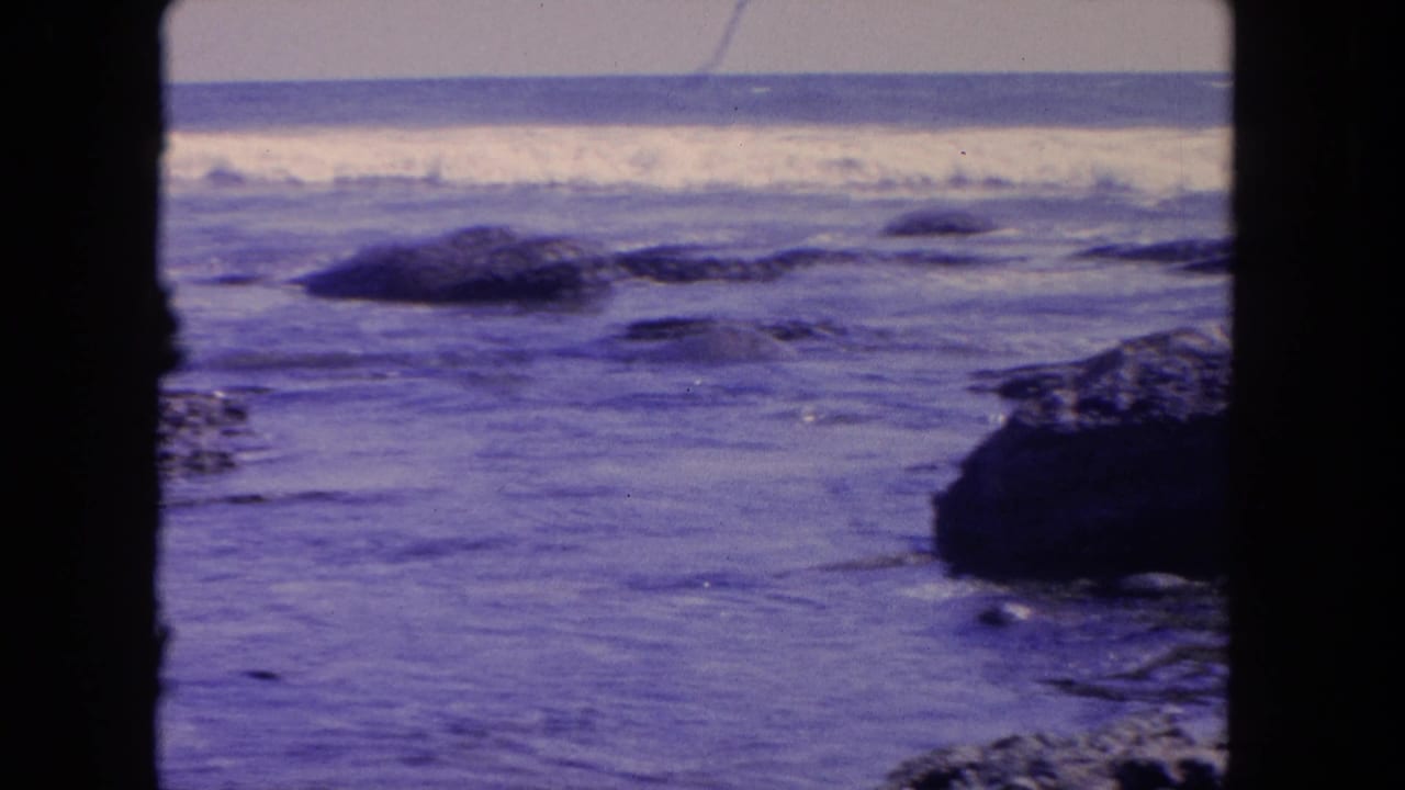 Ocean surf breaks beyond scattered rocks, with a dark vertical frame divider at left.