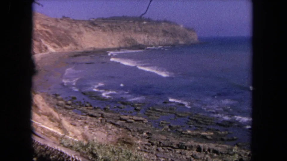 1962 Tijuana coastal wide shot showing cliffs above a sandy beach, rocky shoreline, and small white waves along the bay.