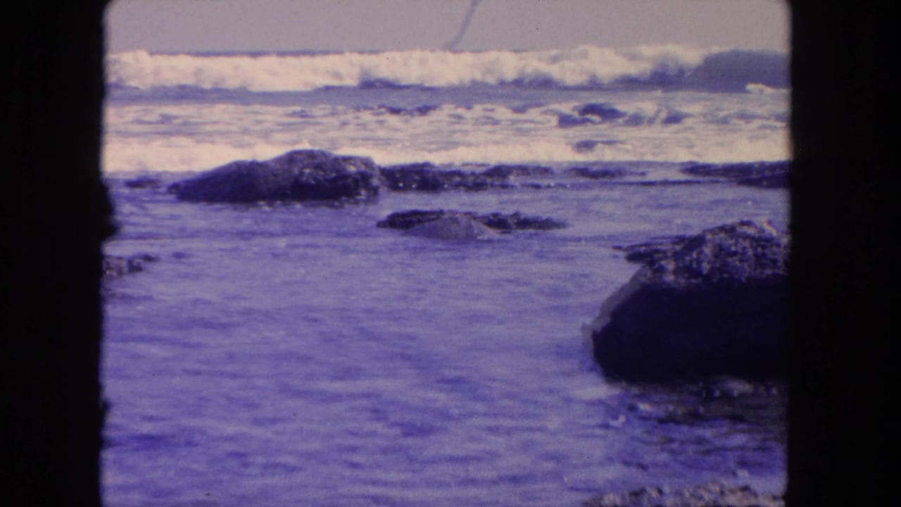 1962 8mm Tijuana coastal view showing surf rolling behind dark shoreline rocks, with blue-purple aged-film color and visible frame edges.