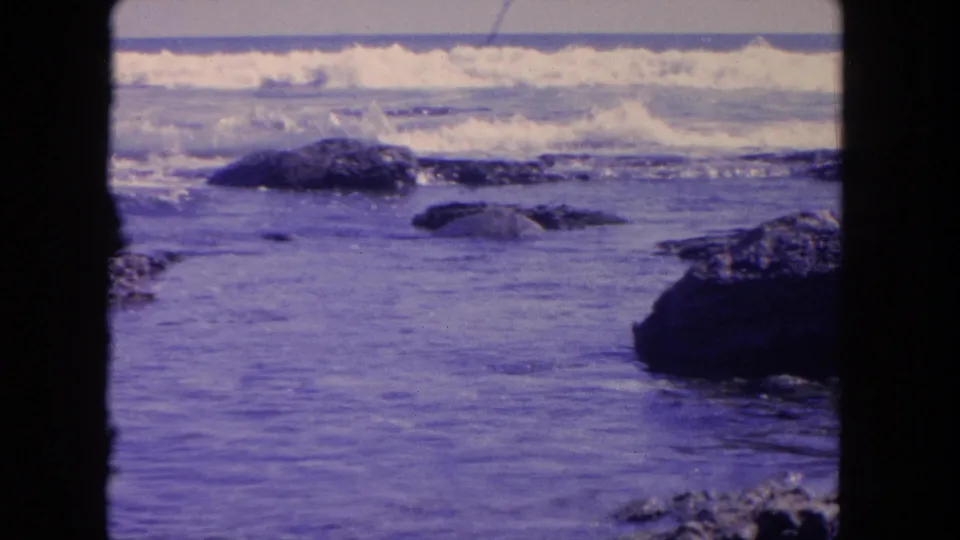 Tijuana coast in 1962 home-movie footage, with foamy surf breaking behind jagged rocks and aged 8mm color visible in the frame.