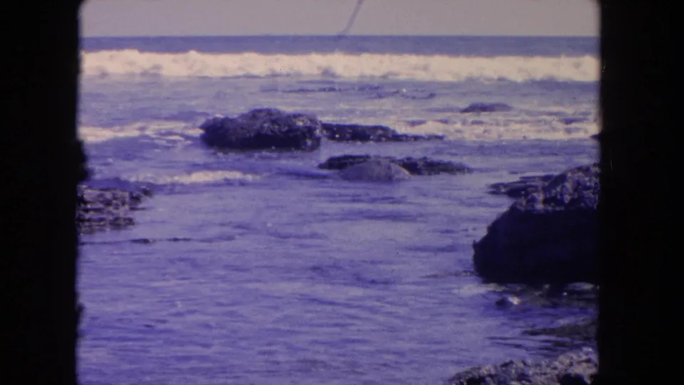 1962 8mm Tijuana shoreline view with dark rocks in the foreground water and white surf breaking across the background.
