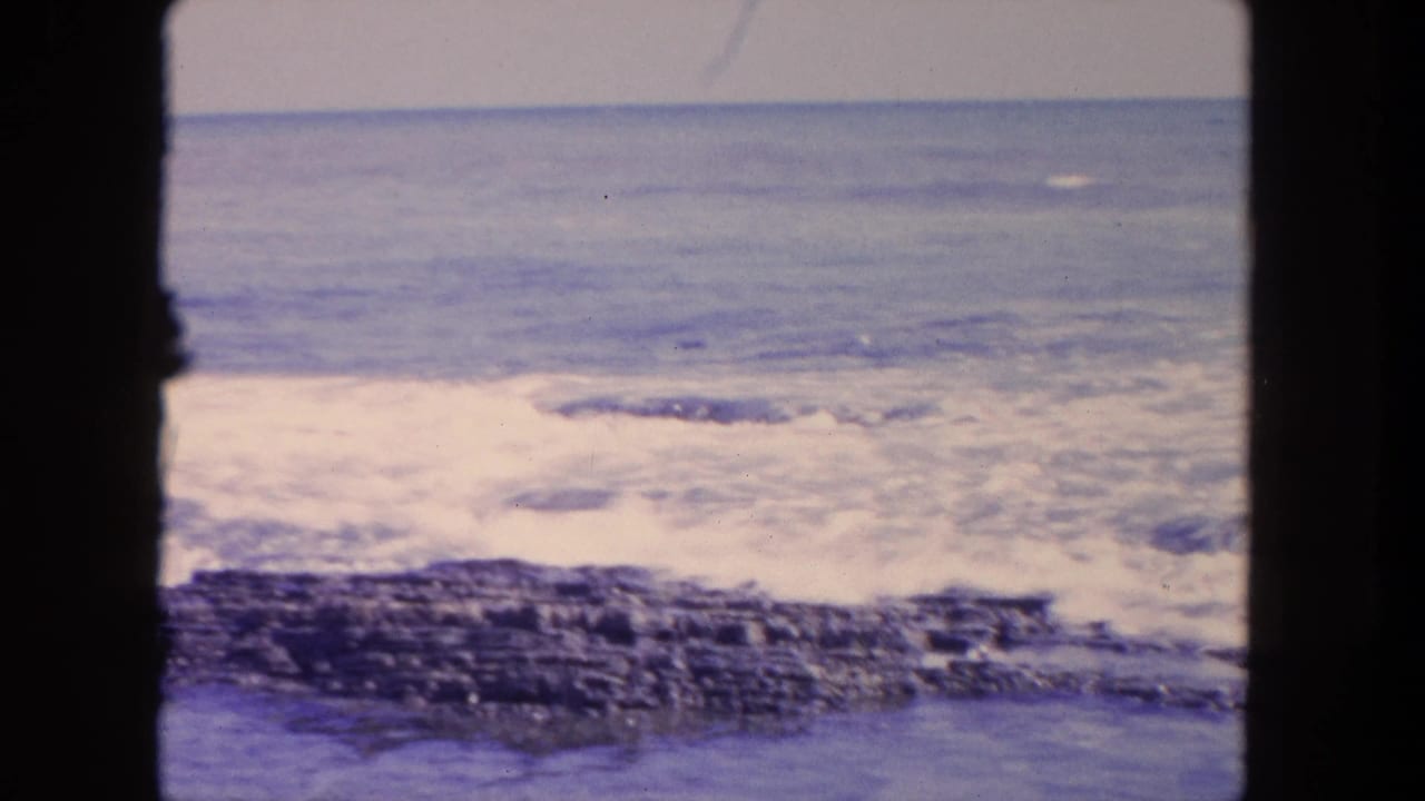 Small waves break over dark rocks beneath a blue ocean horizon.