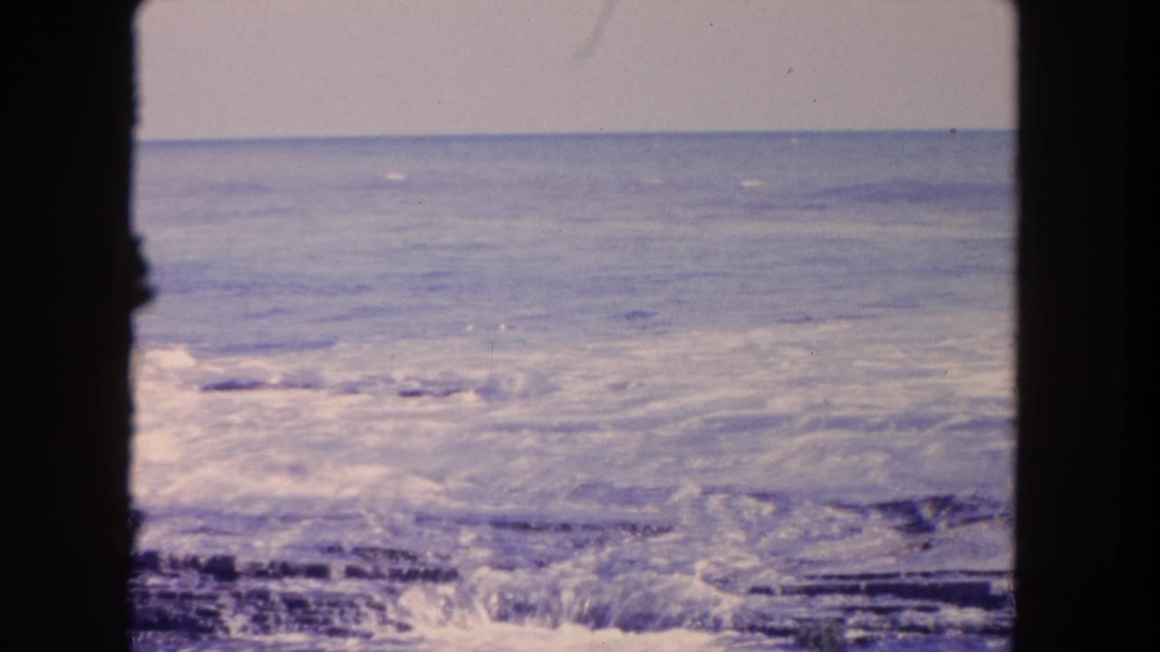 1962 Tijuana coastal frame with foamy surf moving over jagged rocks, seen through dark vertical film borders.