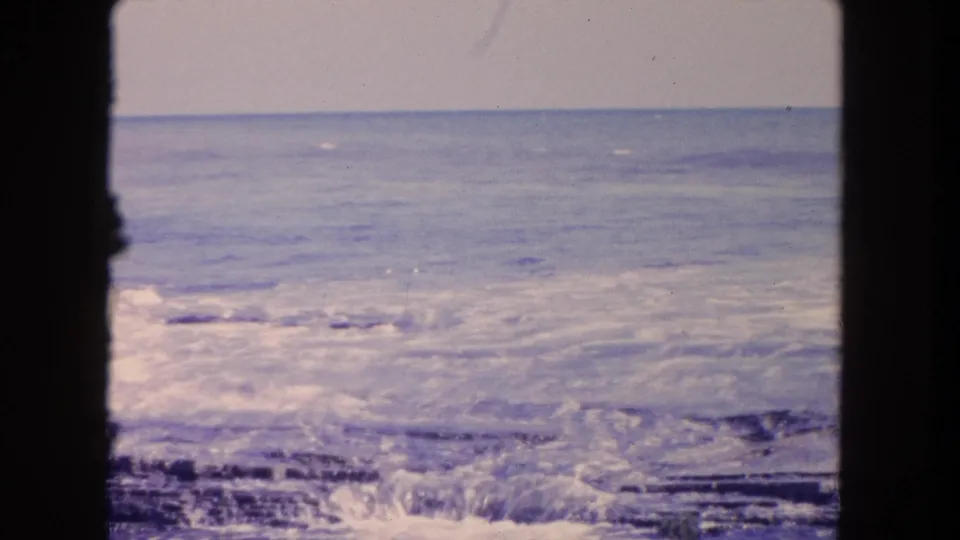 1962 Tijuana coastal frame with foamy surf moving over jagged rocks, seen through dark vertical film borders.