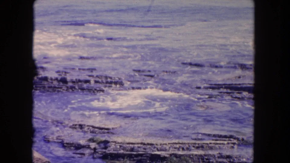 1962 Tijuana 8mm view of shallow water and dark rock shelves with soft purple-blue aging across the frame.