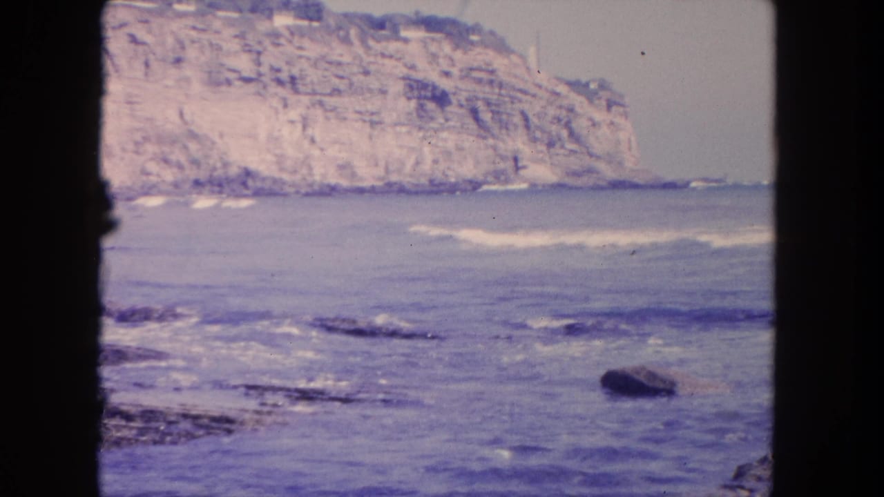 1962 8mm view near Tijuana of blue-purple surf, exposed rocks, and a layered cliff seen through dark frame edges.