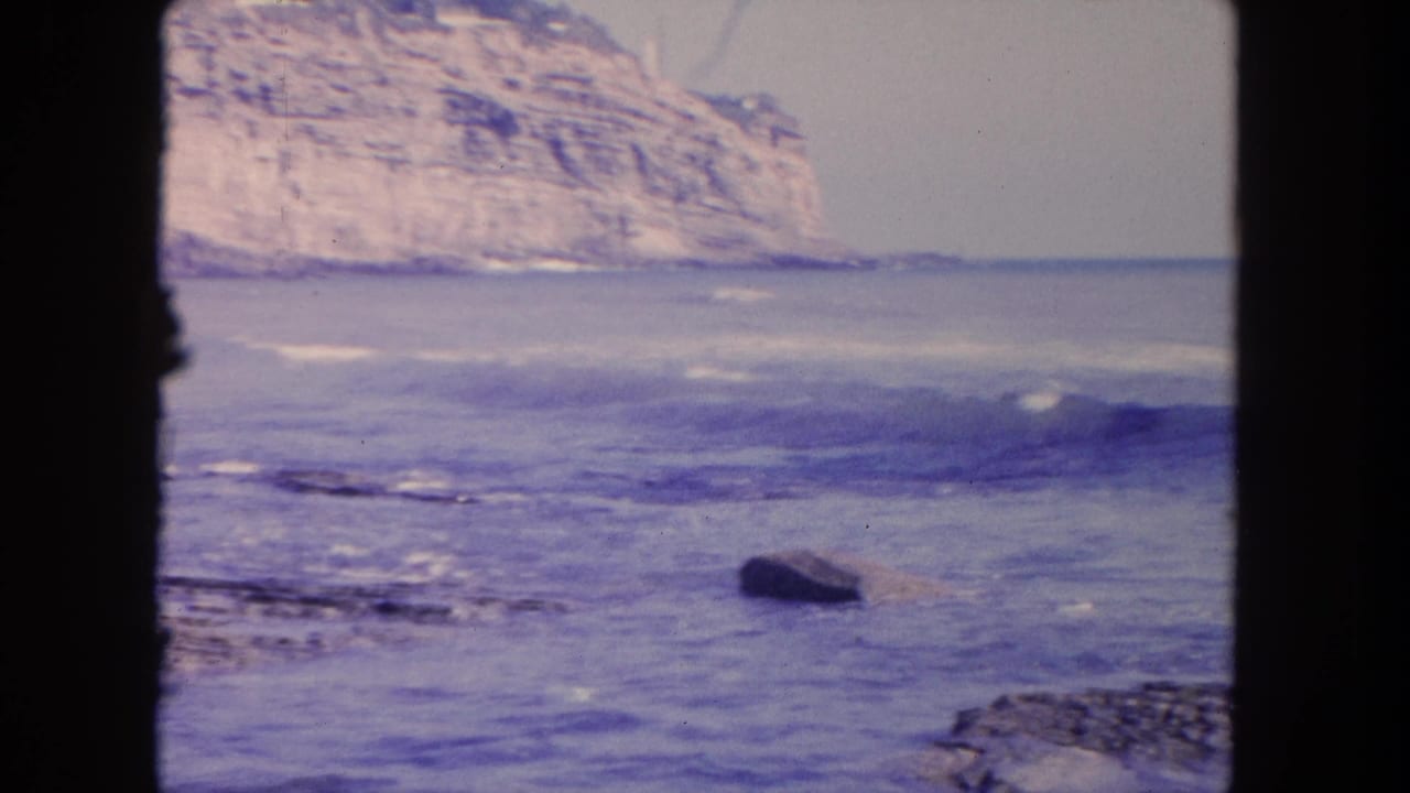 1962 Tijuana coastal frame showing rolling surf, a dark rock in the water, and a pale stratified cliff beyond the shoreline.