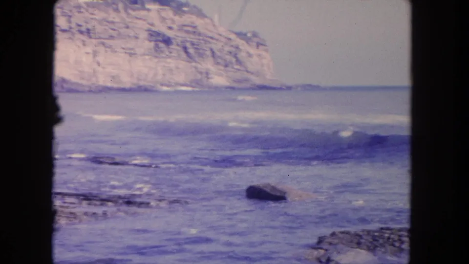1962 Tijuana coastal frame showing rolling surf, a dark rock in the water, and a pale stratified cliff beyond the shoreline.