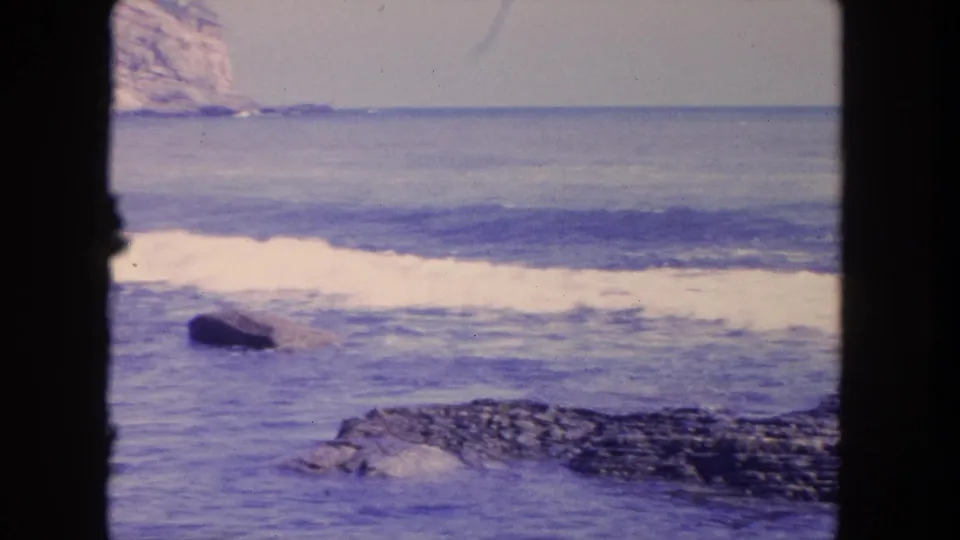 Vintage 8mm frame from the Tijuana coast showing foamy waves, dark tide rocks, and the edge of a cliff at left.