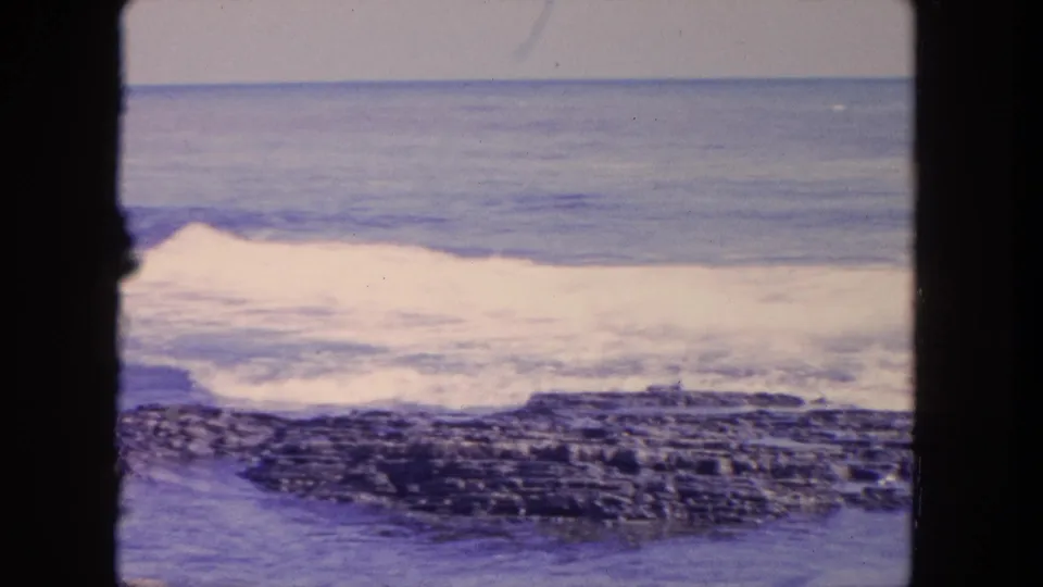 1962 home-movie frame near Tijuana with white surf spreading over exposed rocks and a calm blue horizon beyond.