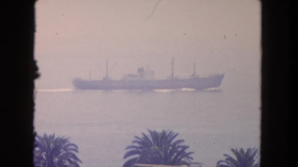 1962 Mexico travel footage shows a distant cargo ship offshore, seen through dark framing with palm tops in the foreground.