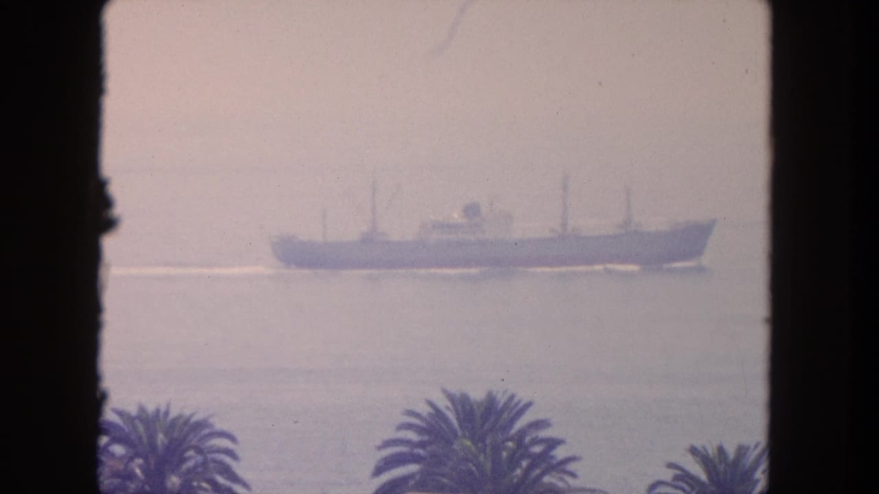 A distant cargo ship passes offshore in hazy 1962 Mexico home-movie footage, with palm tops and a dark window frame visible.