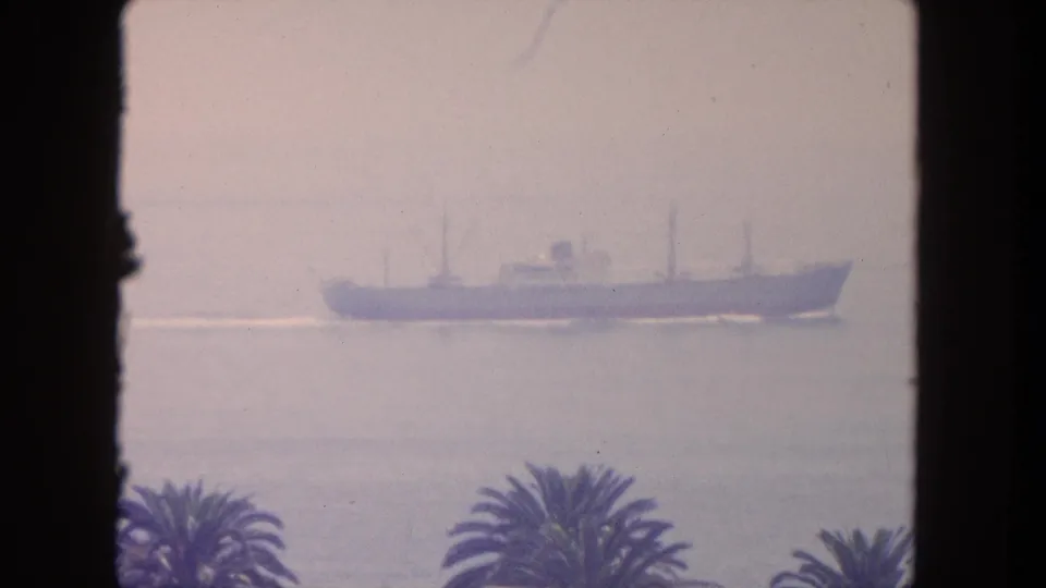 A distant cargo ship passes offshore in hazy 1962 Mexico home-movie footage, with palm tops and a dark window frame visible.