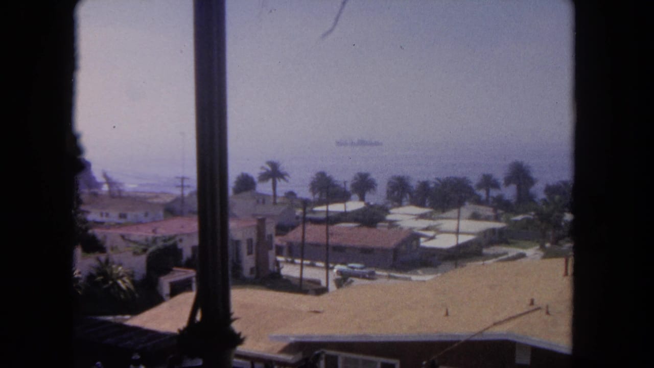 1962 Tijuana coastal view from a window, with palms, low houses, terracotta roofs, ocean haze, and a distant ship offshore.