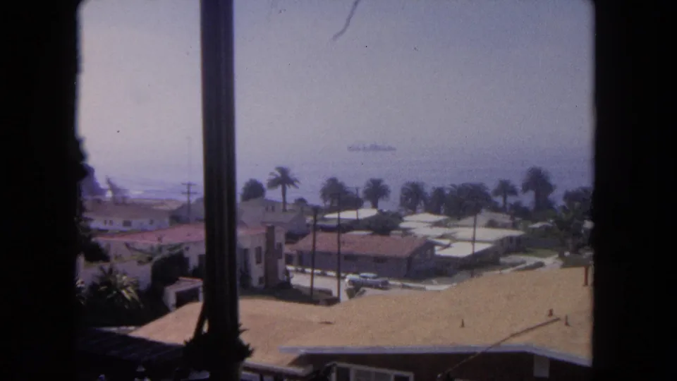1962 Tijuana coastal view from a window, with palms, low houses, terracotta roofs, ocean haze, and a distant ship offshore.