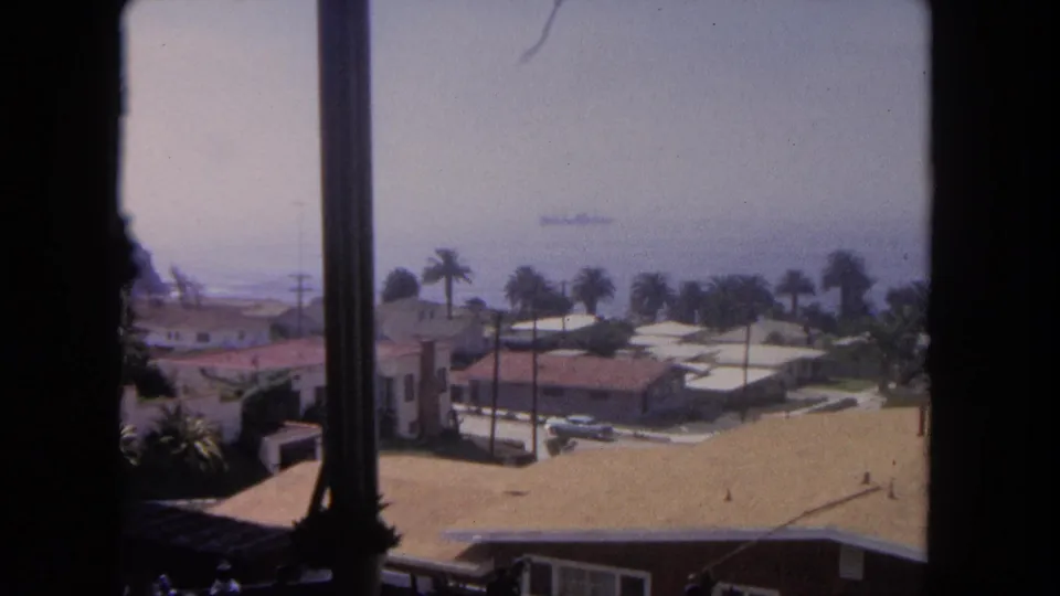 A steady 1962 Tijuana window view shows sunlit rooftops, palm trees, ocean haze, and a distant large ship on the water.