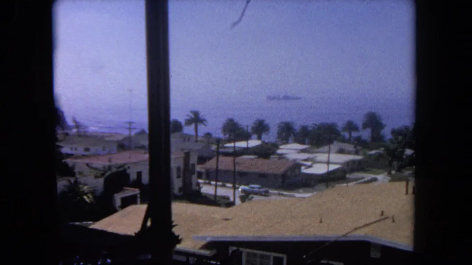1962 Tijuana coastal neighborhood view through a window, with low houses, palm trees, hazy ocean, and a distant ship.