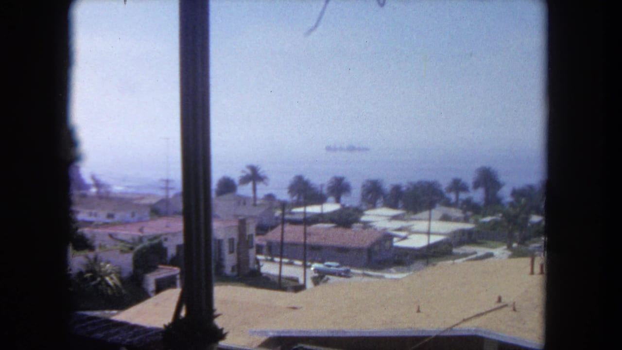 Window-framed view of houses, palms, parked cars, hazy coastline, and a distant ship at sea.